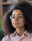 Woman with natural curly hair and glasses meditating indoors with eyes closed and calm expression.