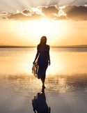 Silhouette of a woman walking through shallow water at sunset, with sun rays breaking through clouds.