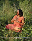A woman in an orange dress sits in meditation with hands on her chest, surrounded by lush green foliage.