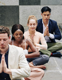 Four professionals sitting in a row outdoors, eyes closed in a meditative prayer pose.