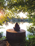 Silhouette of a person meditating on a concrete structure by a calm river under green trees.