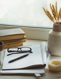 Notebook, books, glasses, pen, and coffee arranged on a desk by a window.