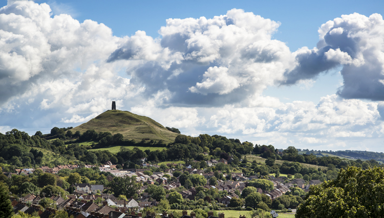 View of Glastonbury Tor on Summer day with partially blue sky and puffy clouds in the background.
