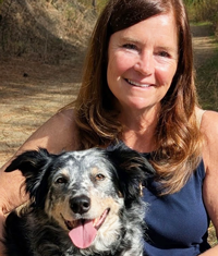 Smiling woman with long brown hair poses with a speckled black-and-white dog outdoors with forest road in background.
