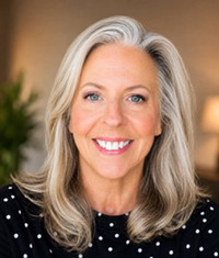 Smiling woman with long gray hair wearing a dark patterned top, seated indoors against a blurred muted background.