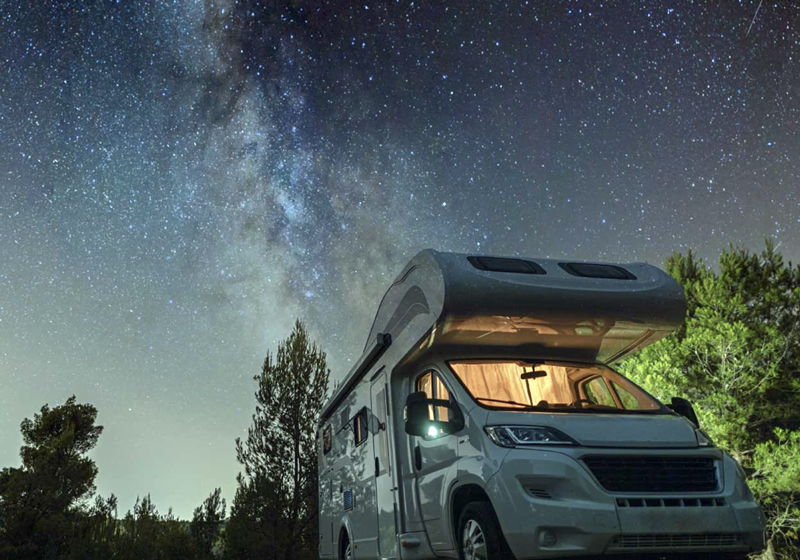 RV parking: White RV parked beneath a star-filled night sky, with the Milky Way visible above trees.
