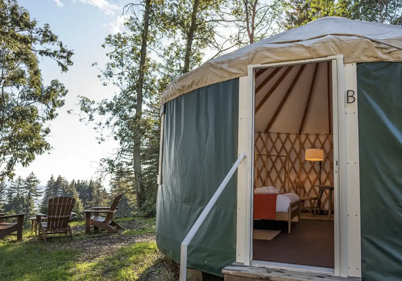 Yurt: Round yurt with an open doorway, bed inside, and Adirondack chairs overlooking a wooded landscape.