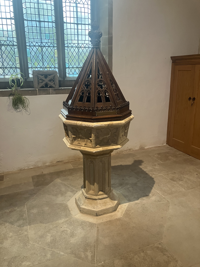 Ornate stone baptismal font with a carved wooden Gothic canopy, beside leaded-glass windows in a church.