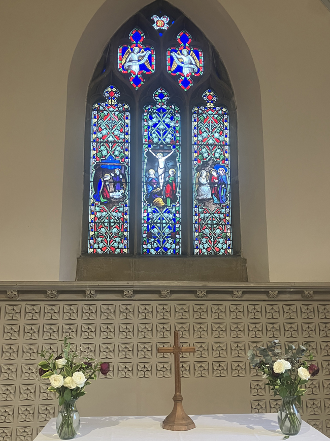 Stained-glass church window depicting the Crucifixion, above an altar with a cross and flower arrangements.