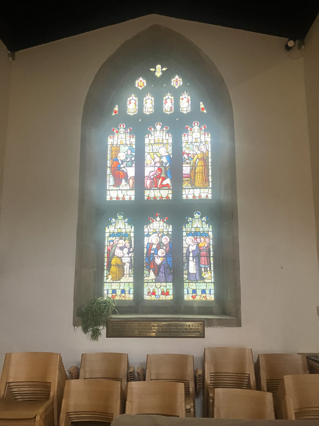 Stained-glass church window depicting biblical scenes, above rows of wooden chairs in the nave.