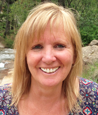 Smiling woman with blonde hair, outdoors by a stream and greenery.
