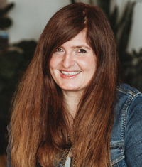 Smiling woman with red hair wearing a jeans jacket, seated indoors against a blurred background.