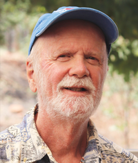 Smiling older man with white beard wearing a blue cap and patterned shirt outdoors.