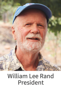 Smiling older man with white beard wearing a blue cap and patterned shirt outdoors.