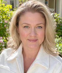 Smiling woman with blond hair wearing a white shirt outdoors with flower bushes blurred in the background.